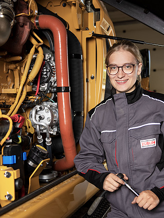 Nahaufnahme einer technischen Maschine, an der eine junge Frau mit Brille lehnt.