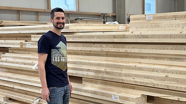 Foto eines jungen Mannes, der in der Produktion vor einem Holzplattenstapel steht und lächelt