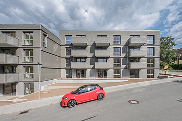 Foto der Joseph-Stiftung in Bayreuth. Man sieht ein Gebäude mit grauer Fassade und ein vorbeifahrendes, rotes Auto.