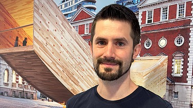 Foto eines Mannes mit dunklen kurzen Haaren und kurzem Bart. Er trägt ein T-Shirt mit Aufdruck. Im Hintergrund sieht man ein Bild mit einer geschwungenen Holzskulptur.